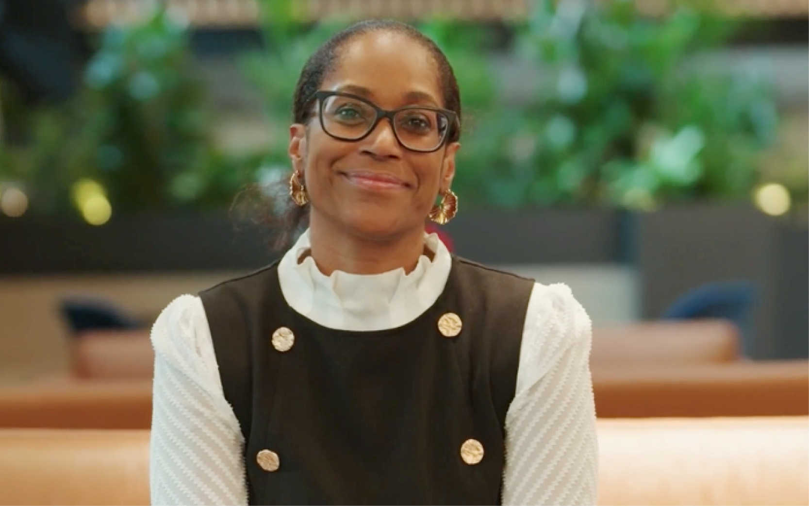Person seated in a modern office lounge, wearing a black sleeveless dress over a white high‑collar textured blouse, accented with gold buttons and gold earrings; soft‑focus greenery and tan seating in the background.